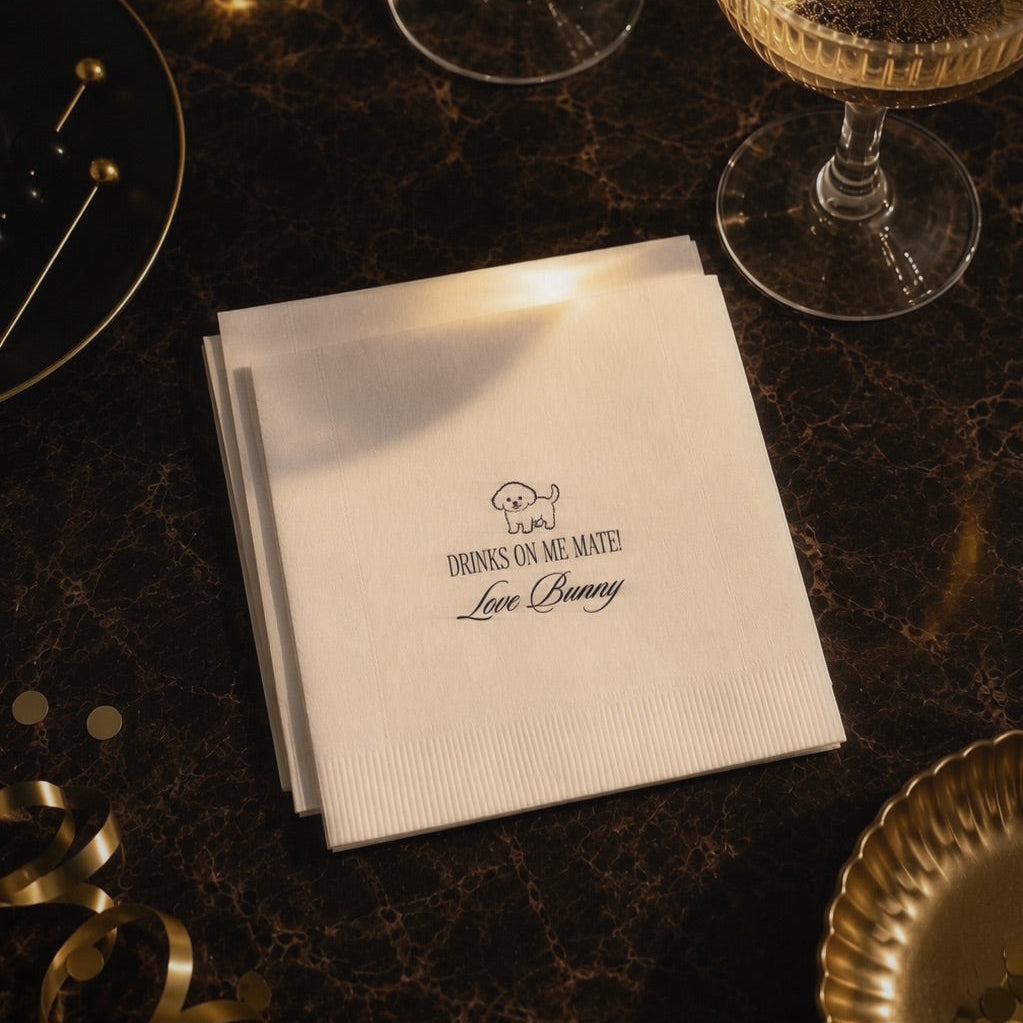 A stack of white paper napkins printed with the phrase 'I'm Drinks on Me Mate! Love Bunny' in black script, styled on a dark marble tabletop with gold-toned glasses, plates, and ribbon.