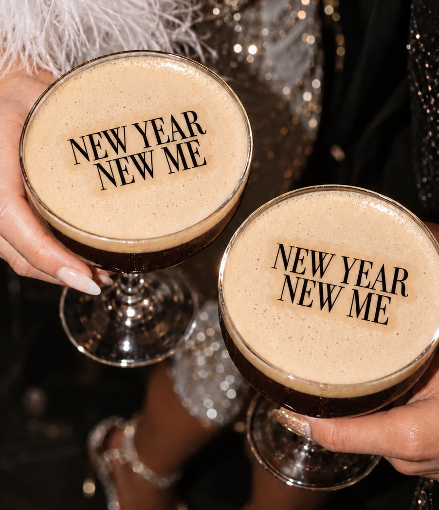 Two hands holding sparkling New Year's Eve cocktails in coupe glasses, with the text 'NEW YEAR NEW ME' printed on the foam. The background shows a person in a sparkling silver embellished dress and silver high-heeled sandals.