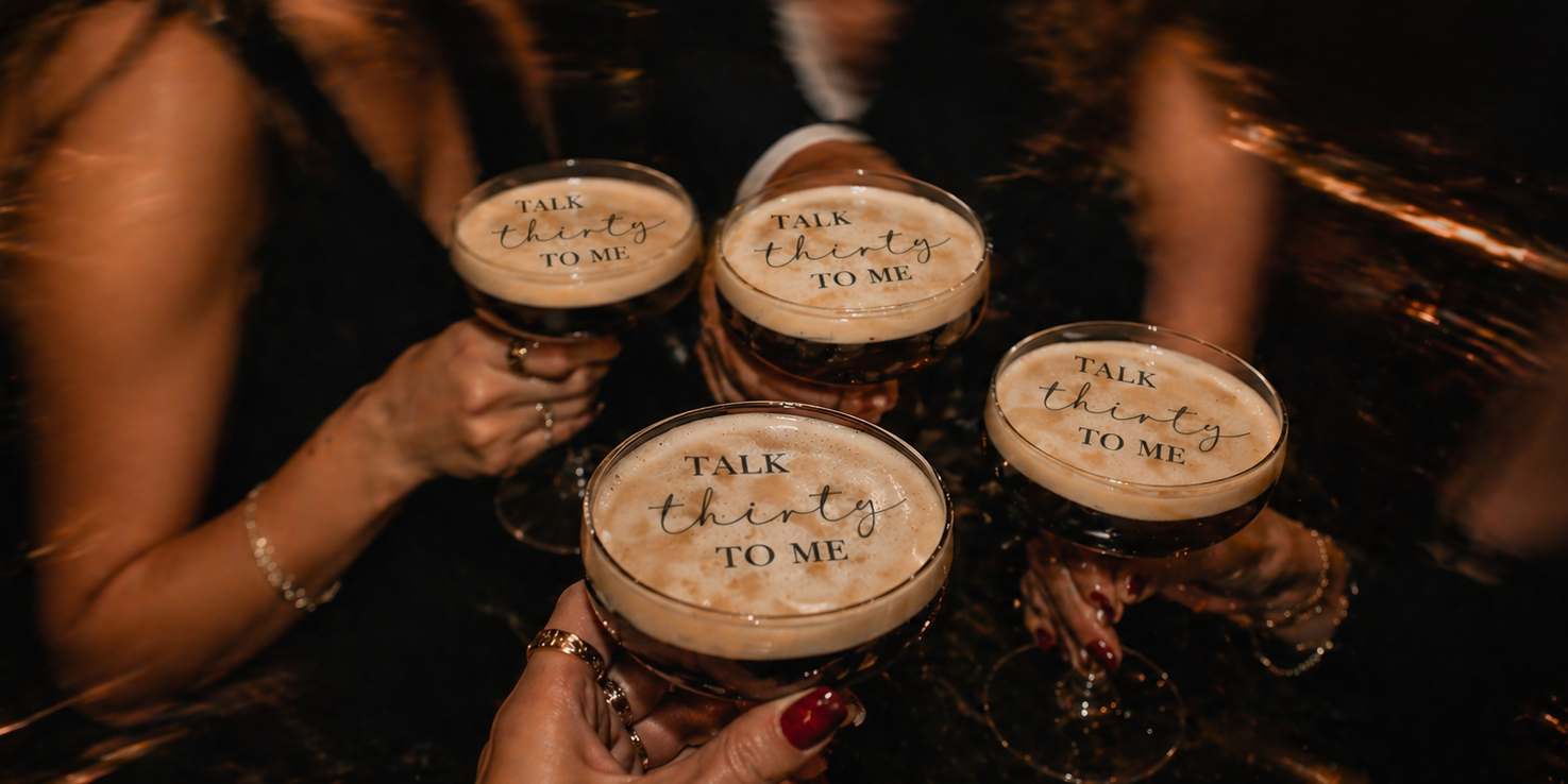 Four cocktail glasses with foam and text held by people in a dimly lit setting