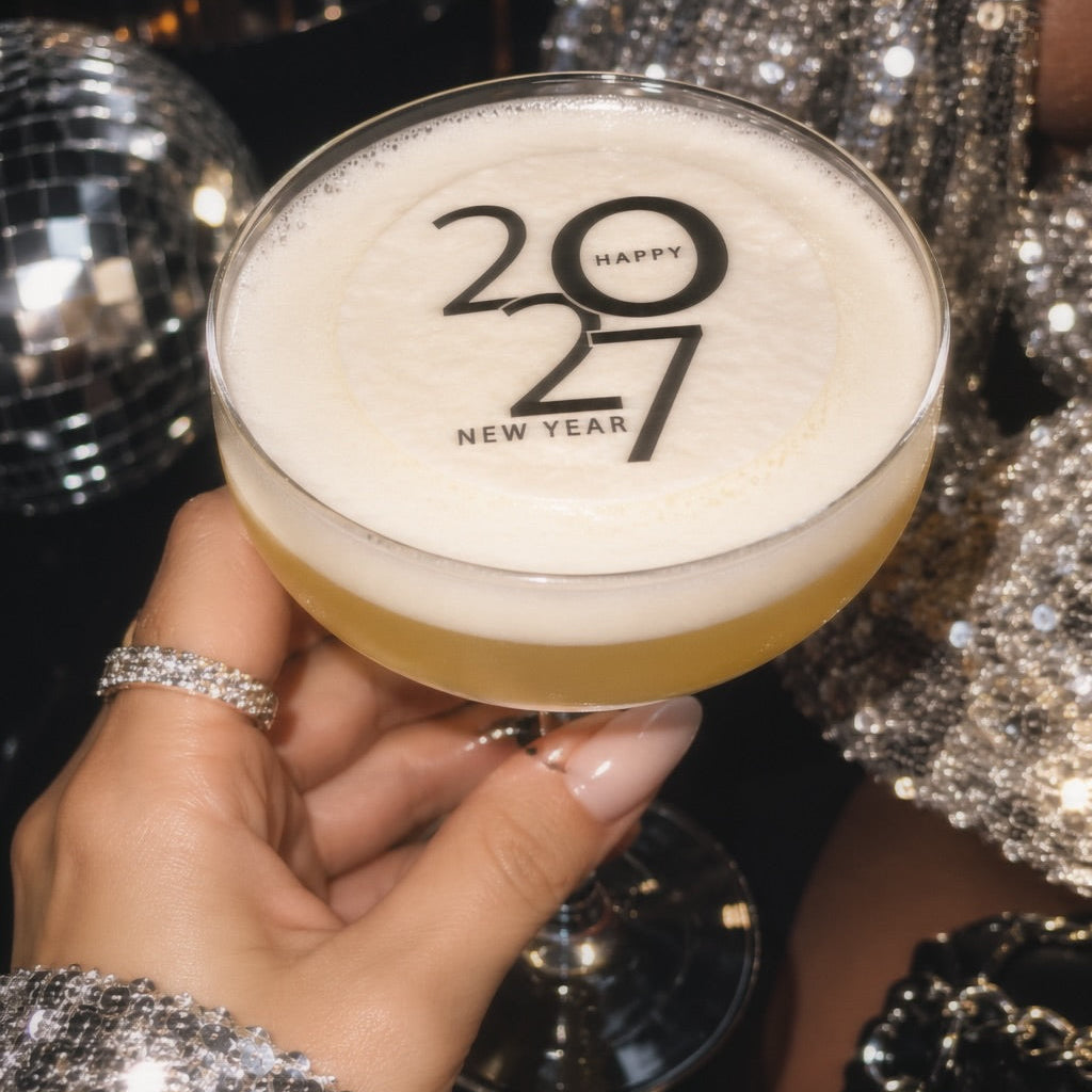 A hand holding a coupe glass filled with a yellow cocktail and thick white foam, printed with '20 Happy 27 New Year' in black. The person is wearing a sparkling silver sequined sleeve and holding the glass in a dark, festive setting with a disco ball in the background.