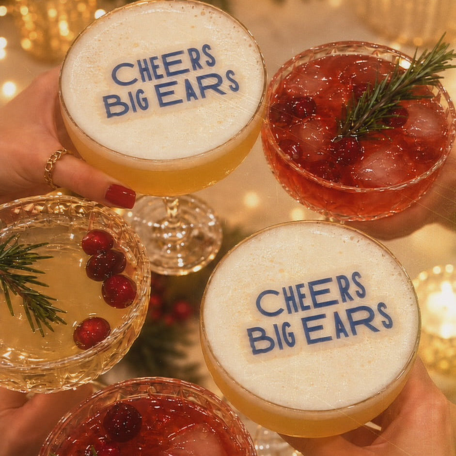 Four festive cocktails in stemmed glasses held by hands, arranged in a toast. The two main drinks have thick white foam tops printed with the blue text 'CHEERS BIG EARS', while the other two contain red berry cocktails with cranberries and rosemary garnish.