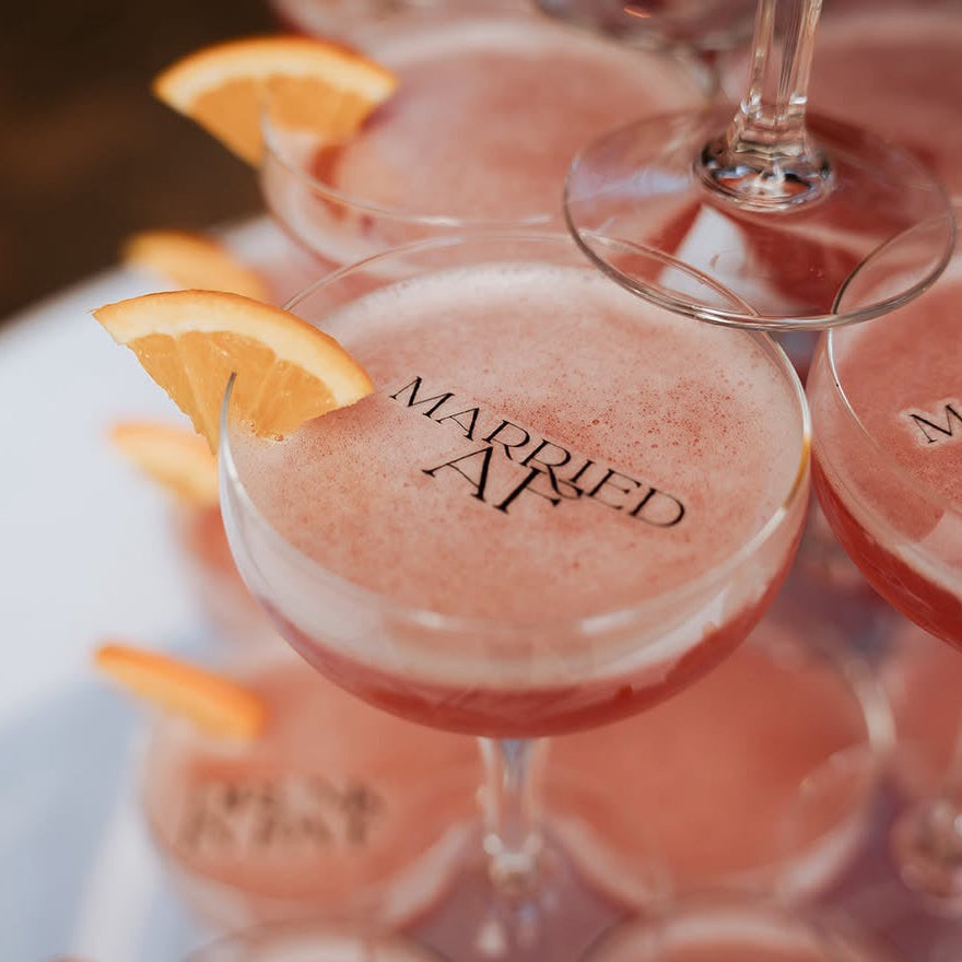 A close-up of several stemmed coupe glasses filled with a pink cocktail and garnished with orange slices, arranged on a white surface. The glasses are printed with the words 'MARRIED AF'.