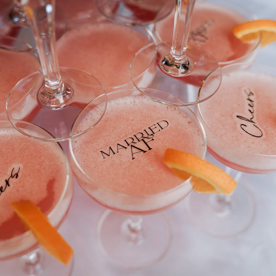 Several coupe glasses filled with a pink cocktail or drink, each garnished with an orange slice. Some glasses have printed text such as 'MARRIED AF', 'Cheers', and 'Chari'.