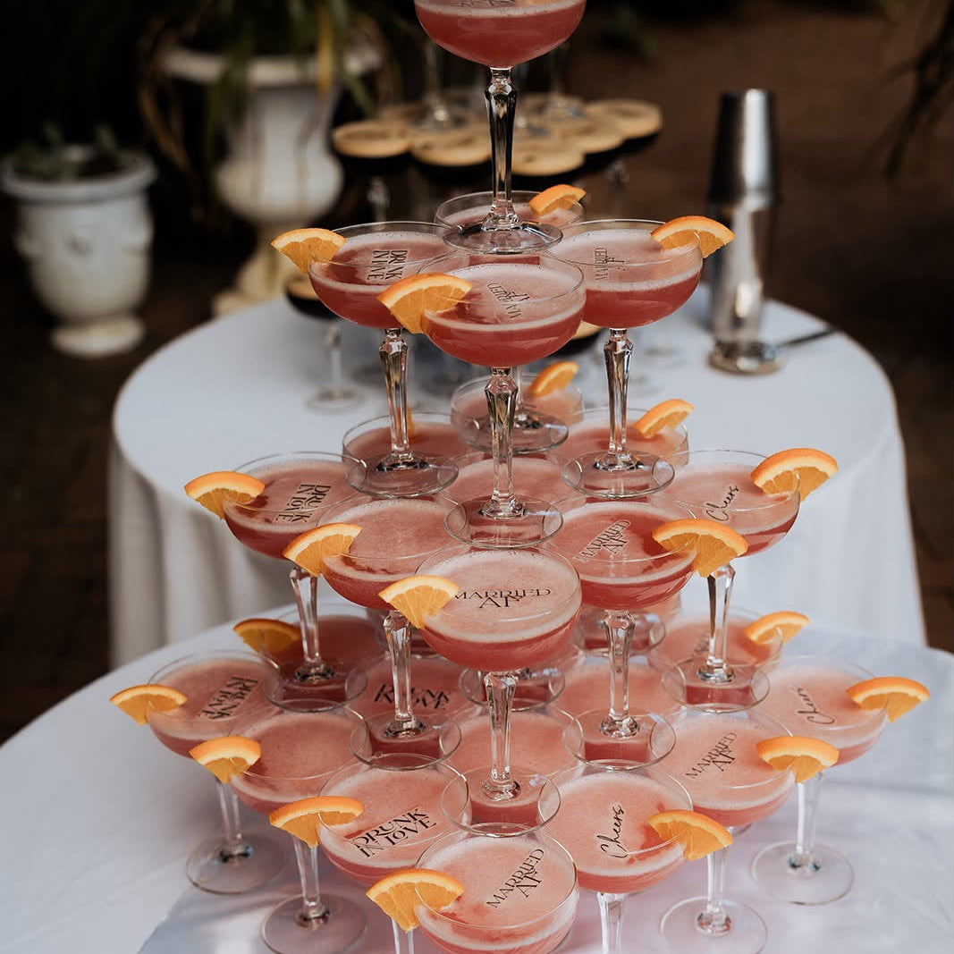 A champagne tower made of stacked coupe glasses filled with pink cocktail sauce, garnished with orange slices, arranged on a white draped table outdoors.