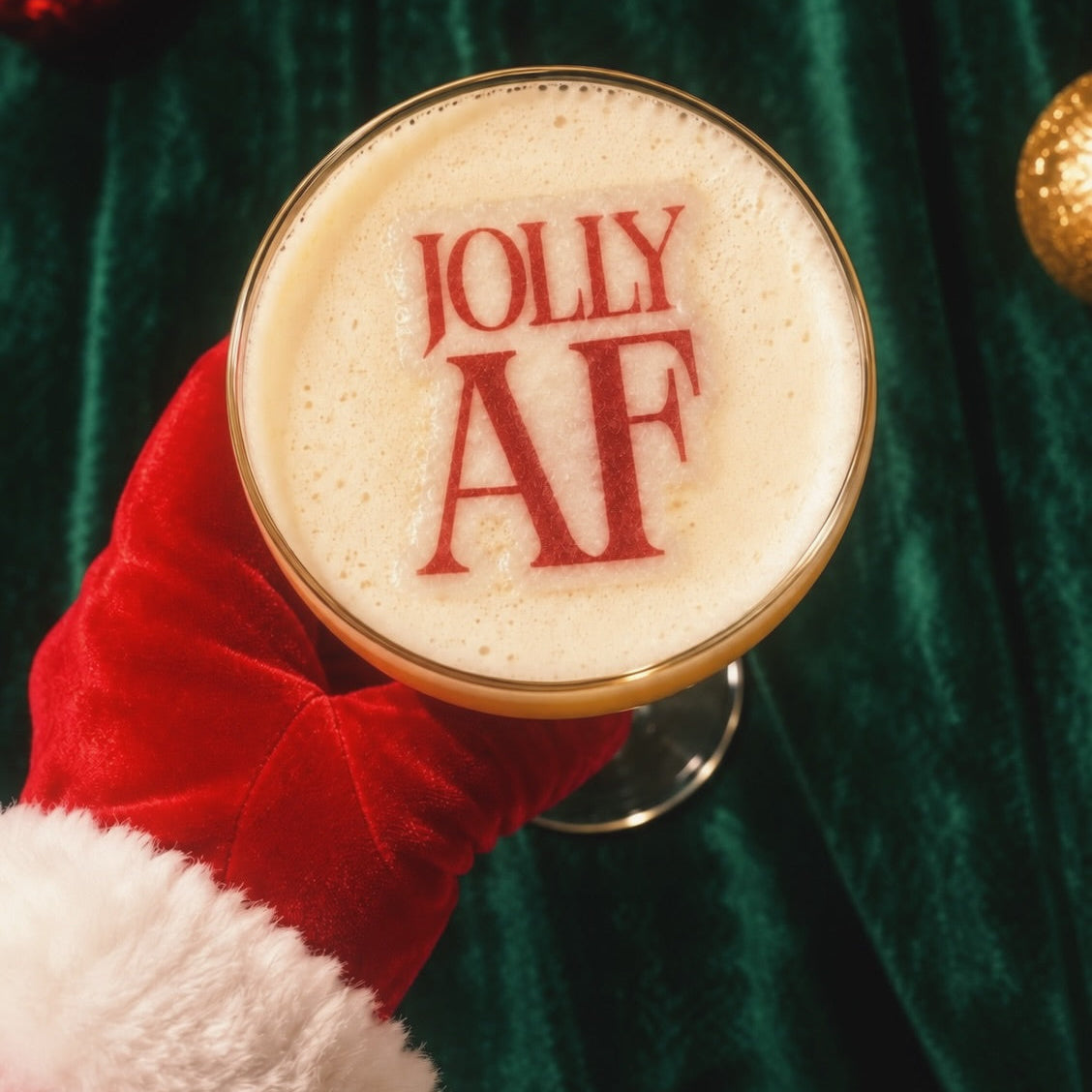 A festive holiday drink in a coupe glass topped with thick foam printed with the words 'JOLLY AF' in red. The glass is held by a hand wearing a red velvet Santa hat glove against a dark green velvet background with Christmas ornaments.