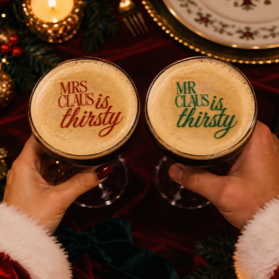 Two hands in Santa sleeves hold glasses of dark stout beer with frothy foam tops printed with holiday quotes: 'MRS CLAUIS is thirsty' in red and 'MR CLAUUS is thirsty' in green. The scene is decorated with candles, greenery, plates, and red velvet fabric.