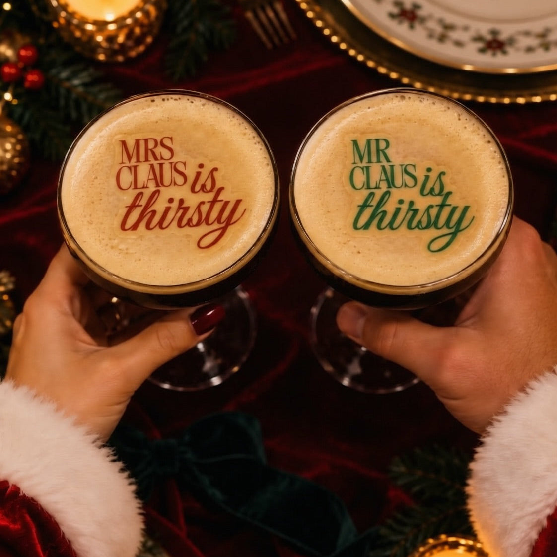 Two hands in Santa sleeves hold glasses of dark stout beer with frothy foam tops printed with holiday quotes: 'MRS CLAUIS is thirsty' in red and 'MR CLAUUS is thirsty' in green. The scene is decorated with candles, greenery, plates, and red velvet fabric.