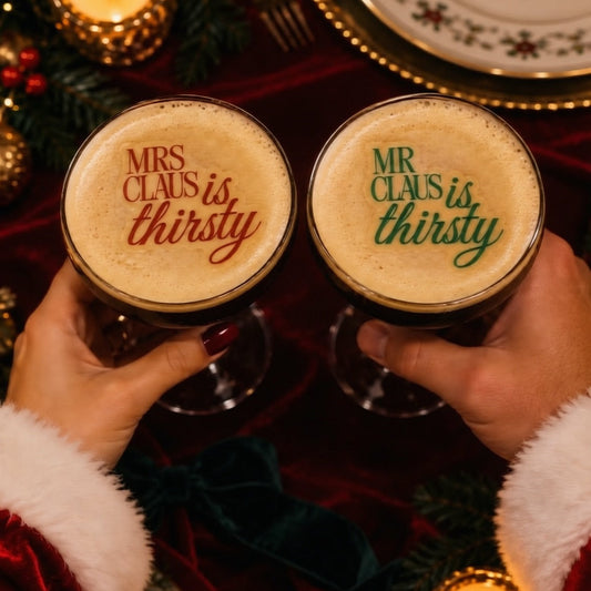 Two hands in Santa sleeves hold glasses of dark stout beer with frothy foam tops printed with holiday quotes: 'MRS CLAUIS is thirsty' in red and 'MR CLAUUS is thirsty' in green. The scene is decorated with candles, greenery, plates, and red velvet fabric.