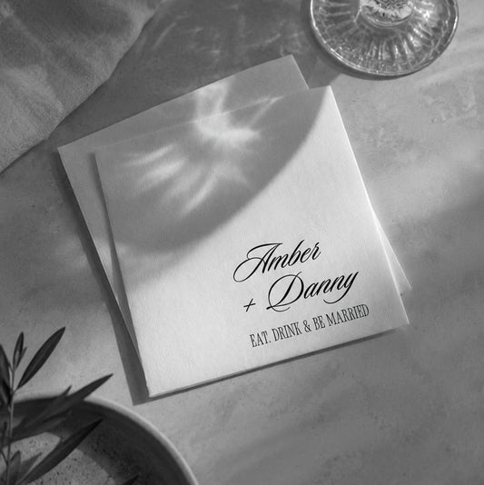 A stack of white paper napkins with elegant black script reading 'Amber + Daney' and 'EAT, DRINK & BE MARRIED' are arranged on a tabletop with soft natural shadows. A glass, draped fabric, and plant leaves are visible around them in a styled setting.