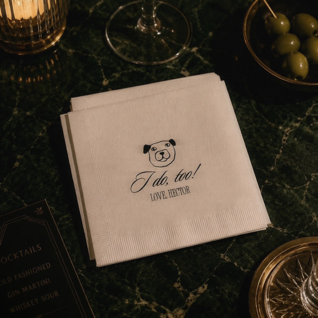 A stack of white paper cocktail napkins on a dark green marble table, printed with a small bear face and the text 'I do, too! LOVE YOU'. The napkins are styled with a gold-rimmed coupe glass, a lit candle in a ribbed holder, and green olives.