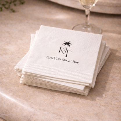 Stack of white napkins with a palm tree design and text on a beige surface with a glass of champagne.