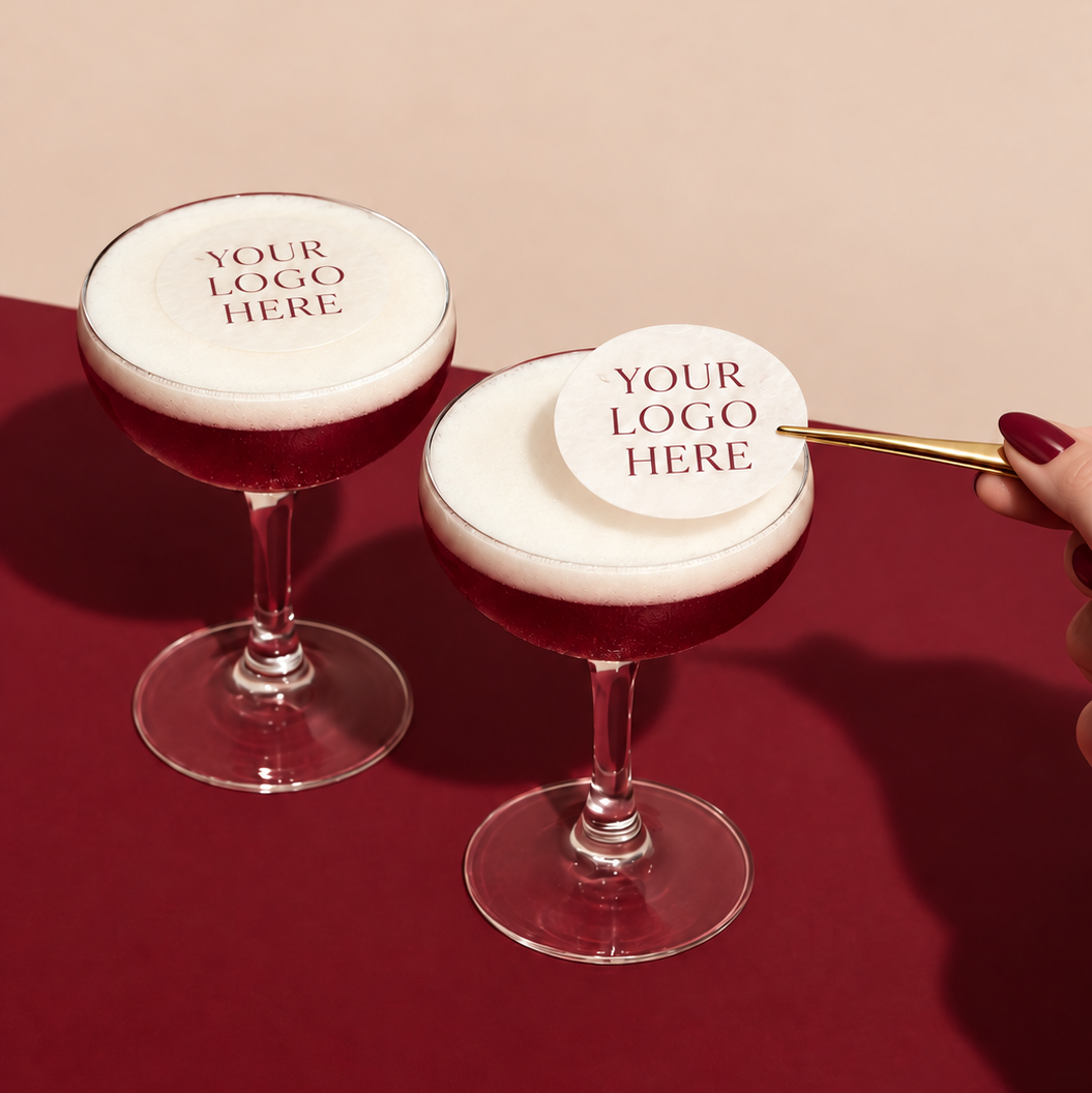 Two stemmed coupe glasses filled with a deep red beverage and topped with thick white foam, each featuring a round white topper printed with placeholder text 'YOUR LOGO HERE.' One topper is placed on the foam of one glass, while a hand with dark red nail polish holds the second topper on a thin gold stick over the foam of the other glass.