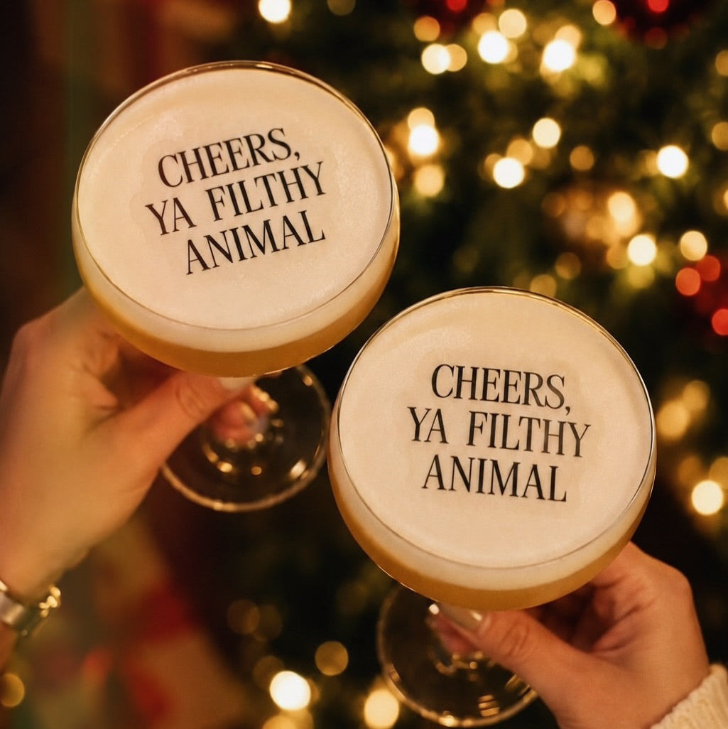 Two hands holding frothy cocktails in coupe glasses with the printed phrase 'CHEERS, YA FILTHY ANIMAL' on the foam, set against a warm, blurred Christmas tree background with bokeh lights.