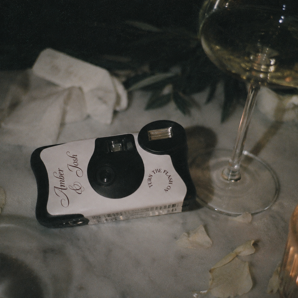 A retro-style instant camera with a white and black body and 'Homer & Jane' printed on the side, resting on a marble tabletop surrounded by a wine glass, napkins, and a candle.