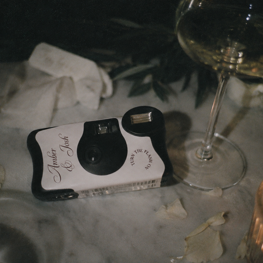 A retro-style instant camera with a white and black body and 'Homer & Jane' printed on the side, resting on a marble tabletop surrounded by a wine glass, napkins, and a candle.