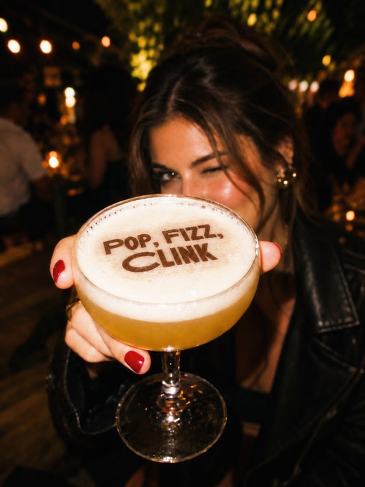 A woman in a black leather jacket holds up a coupe glass filled with an amber cocktail and thick white foam. The foam is printed with the words 'POP, FIZZ, CLINK' in dark brown.